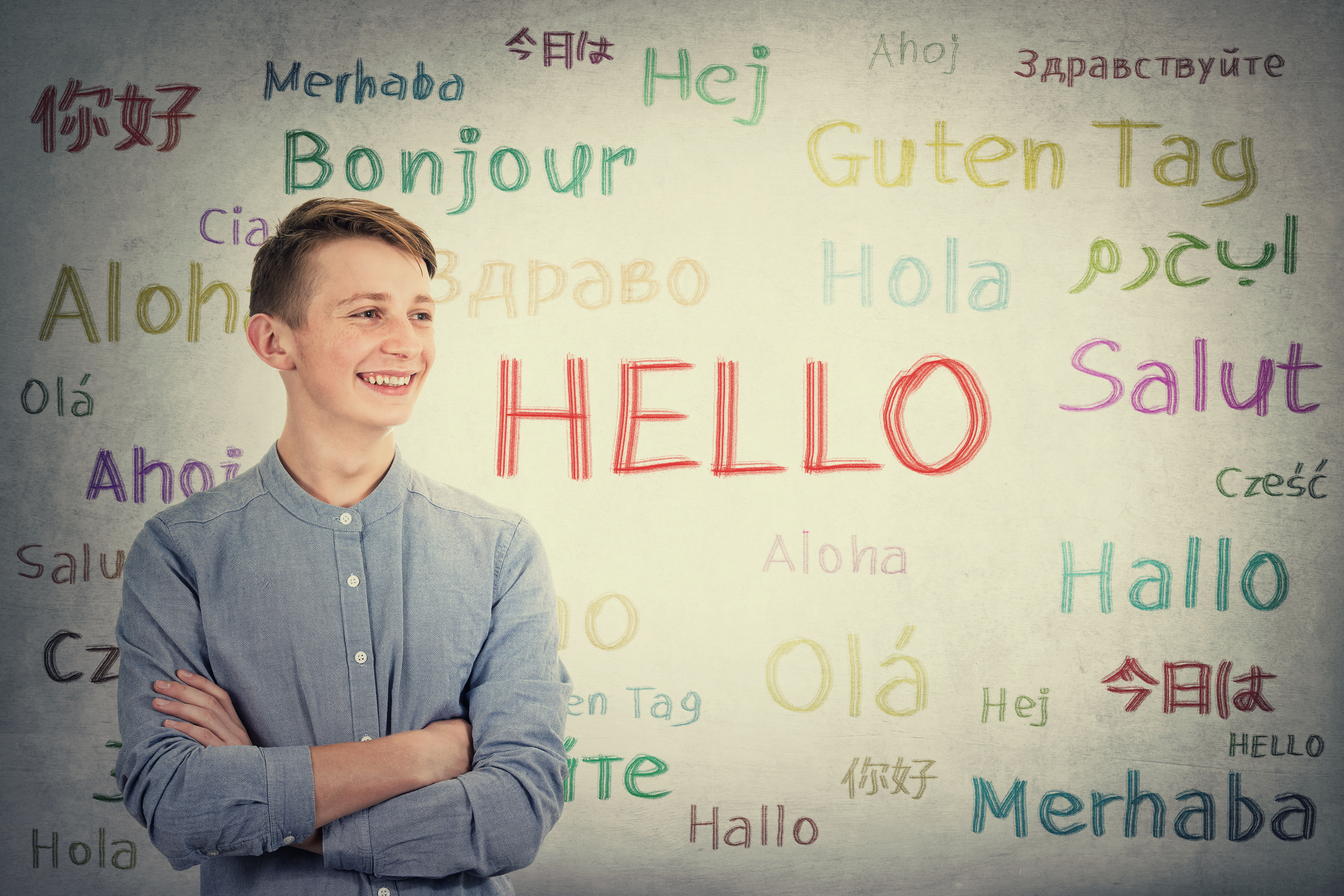 Positive boy adolescent keeps arms crossed looking aside tooth smile over grey wall background written with the word hello in different languages and colors. Learning opportunity for young students.